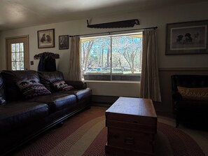 A living room with a leather sofa, a wooden chest, and a window with a view of trees. - Serene 2-bedroom cabin with nestled in the pines. (Pine)