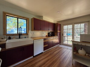 A kitchen with wooden cabinets, a dishwasher, a sink, and a window with a view of trees. - Serene 2-bedroom cabin with nestled in the pines. (Pine)