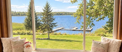 A room with a large window overlooking a lake, a wooden bench with decorative pillows, and a bookshelf with various items.