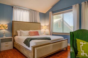Une chambre à coucher avec un lit, des tables de chevet, une fenêtre avec des stores et un fauteuil vert.