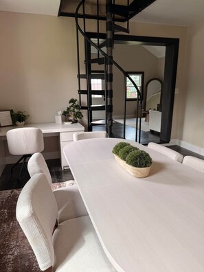 A modern dining area with a white table, chairs, and a central potted plant. A spiral staircase is visible in the background.