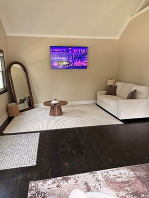 A living room with a dark wooden floor, a white sofa, a round coffee table, a large mirror, and a flat-screen TV mounted on the wall. - Modern Chalet · 8 Min to Holiday Valley · Sleeps 6 (East Otto)