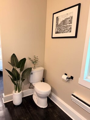 A bathroom with a toilet, a framed black and white photo on the wall, a potted plant, and a toilet paper holder.