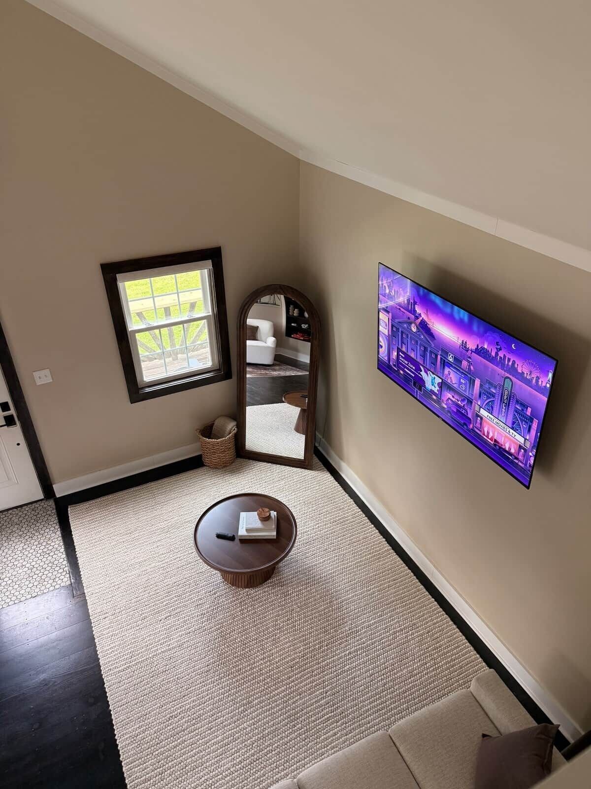 A living room with a flat-screen TV mounted on the wall, a round coffee table with a book, a small rug, and a window with a view of the outdoors.