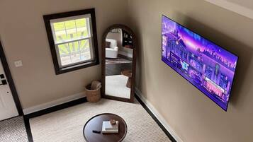 A living room with a flat-screen TV mounted on the wall, a round coffee table with a book, a small rug, and a window with a view of the outdoors.