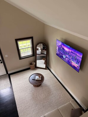 Ein Wohnzimmer mit einem an der Wand befestigten Flachbildfernseher, einem runden Couchtisch mit einem Buch, einem kleinen Teppich und einem Fenster mit Blick ins Freie.
