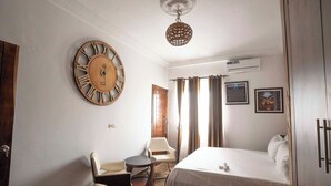 Habitación de hotel con una cama, un reloj de pared redondo, una silla, una mesita, una lámpara colgante y cortinas.