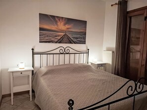 A bedroom with a bed, a nightstand, a bedside table, a lamp, a window with curtains, and a painting of a sunset over the ocean. - Apartment  by Interhome (Lazise)