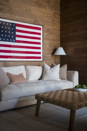 Ein Zimmer mit einem weißen Sofa, einem gewebten Couchtisch, einer Lampe und einer großen amerikanischer Flagge an der Wand.