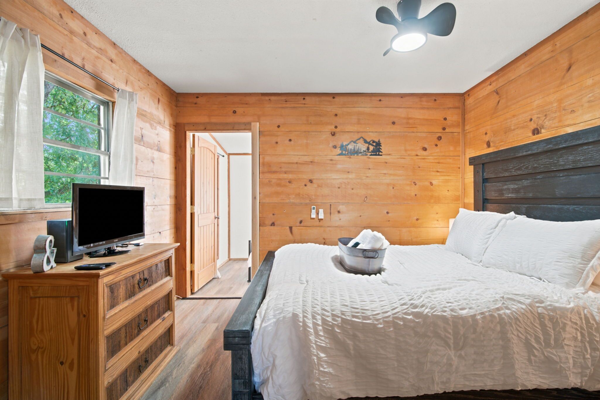 Una habitación con paneles de madera, una cama, un televisor sobre un tocador de madera y una ventana con cortinas.