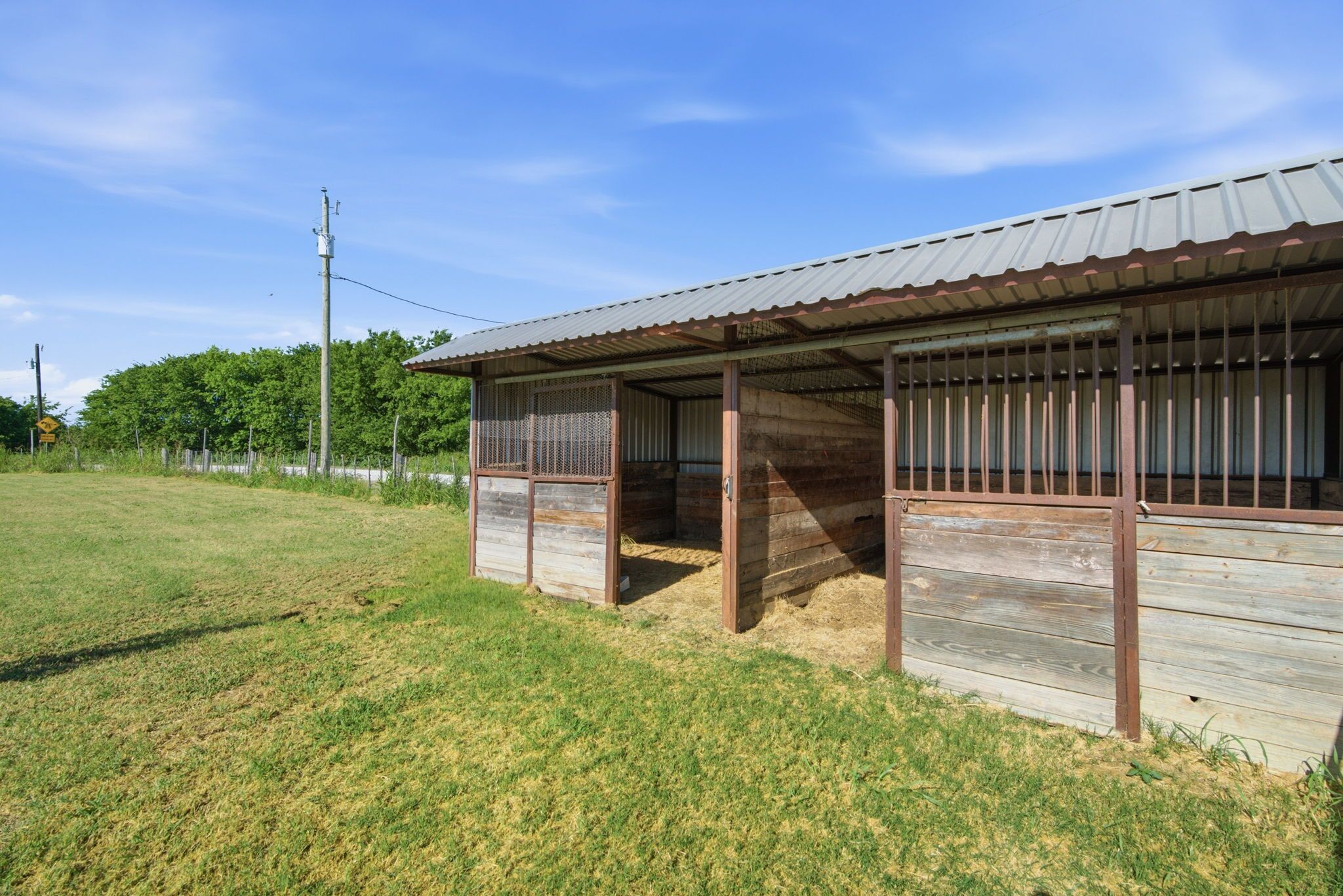 A stable with a metal roof and wooden walls, surrounded by a fenced area and a grassy field.