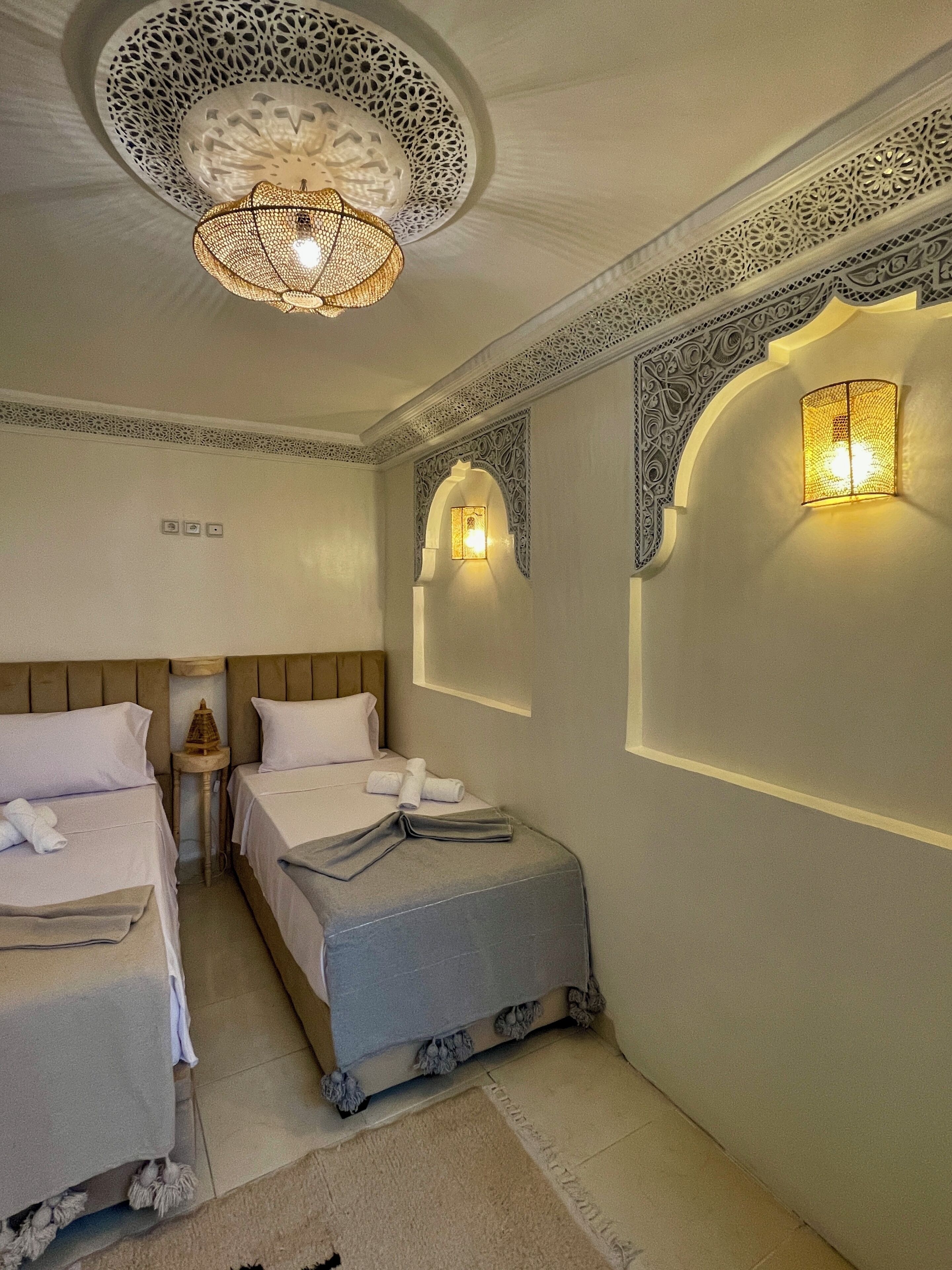 A hotel room with two beds, a decorative ceiling light, wall-mounted sconces, and ornate wall designs.