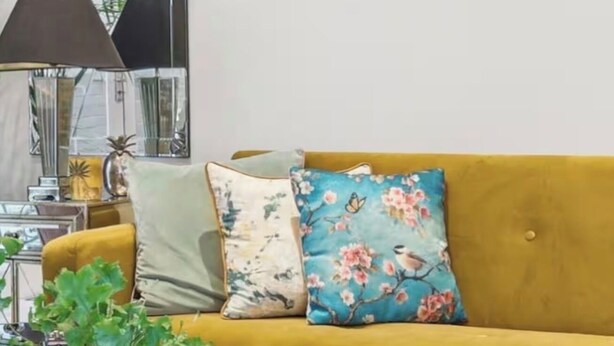 Un salon avec un canapé couleur moutarde, des coussins à motifs floraux, un ventilateur, un miroir et une plante en pot.