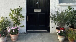 Une porte noire portant le numéro 28, un mur en briques blanches et des plantes en pot disposées au sol.