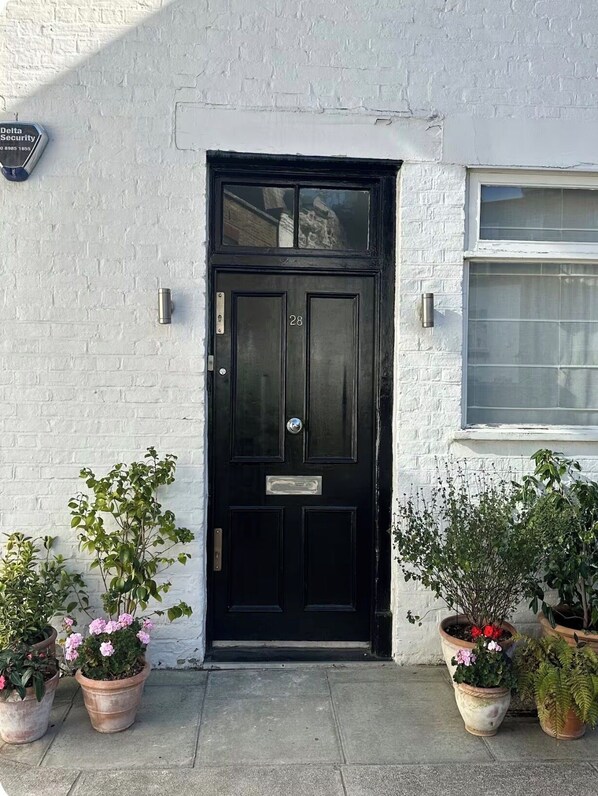 A black door with the number 28, a white brick wall, and potted plants on the ground. - Beautiful Mews house just off Marylebone high st (London)