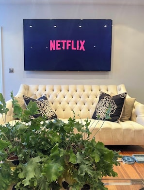 A living room with a tufted sofa, a large flat-screen TV mounted on the wall, and a potted plant in the foreground.