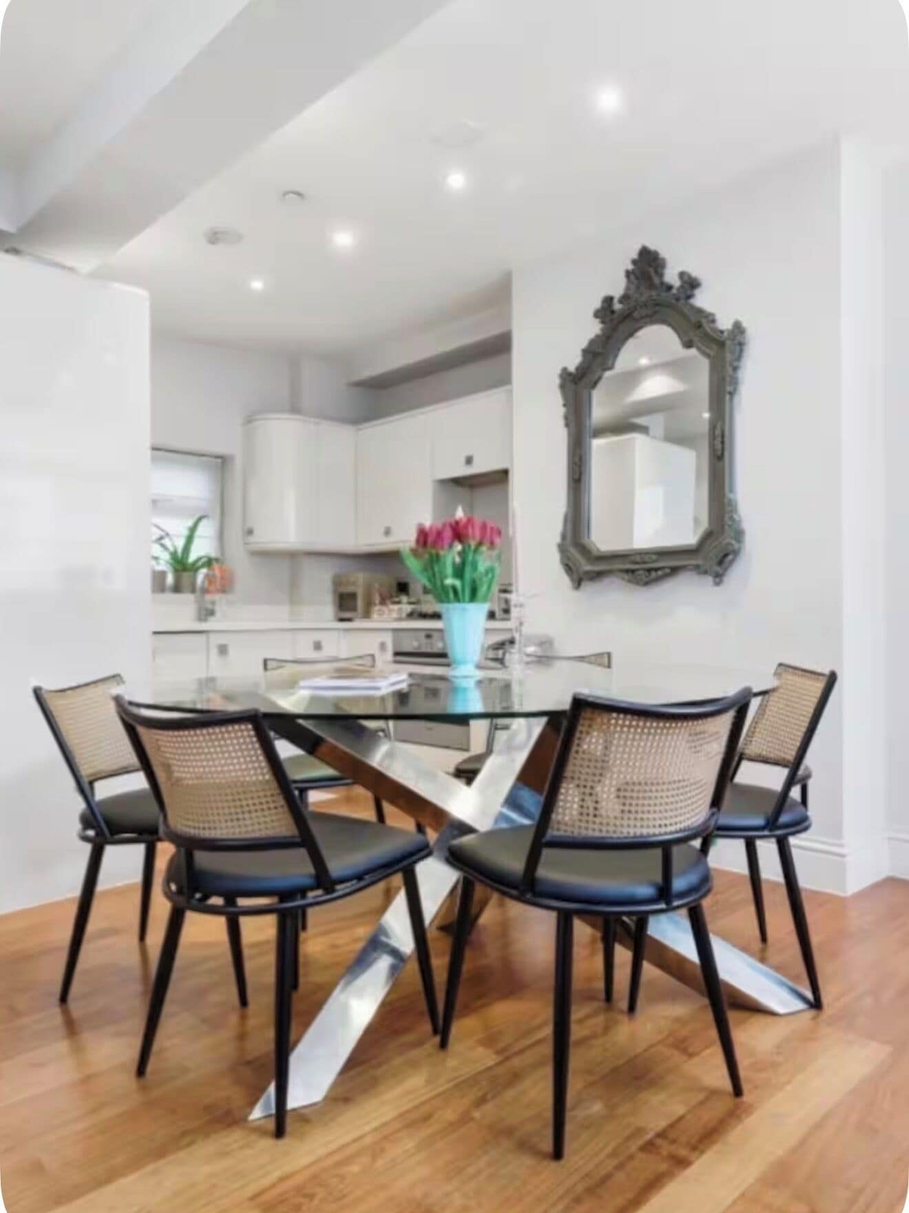 A dining area with a glass table, four chairs, a vase with flowers, and a mirror on the wall.