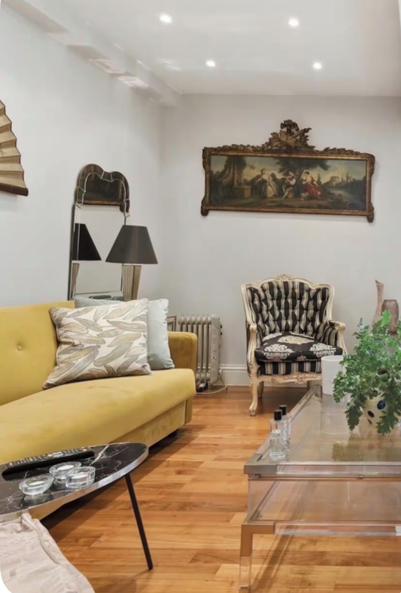 A living room with a yellow sofa, a patterned armchair, a glass coffee table, and a painting on the wall.