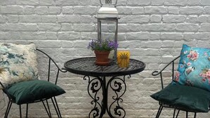 Une table ronde en métal, ornée d’un pot de fleurs et d’une bougie, est flanquée de deux chaises dotées de coussins à motifs floraux, le tout adossé à un mur de briques blanches.