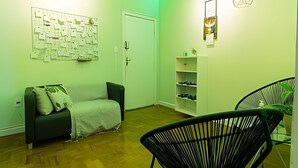 A room with a green wall, a bulletin board, a sofa, a chair, a shelf with shoes, and a hanging light fixture. - Suíte Ampla Augusta Paulista Frei Caneca (São Paulo)