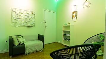 Um quarto com parede verde, um mural, um sofá, uma cadeira, uma estante com sapatos e um candeeiro suspenso.