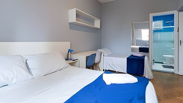 A hotel room with a large bed, a bedside table, a chair, a mirror, and a bathroom visible through a glass door. - Suíte Ampla Augusta Paulista Frei Caneca (São Paulo)