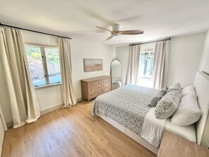Ein Schlafzimmer mit einem Bett, einer Kommode, einem Deckenventilator und zwei mit Vorhängen versehenen Fenstern.