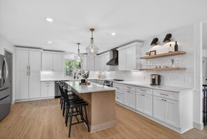 A modern kitchen with white cabinetry, a central island with stools, pendant lighting, and a view of greenery outside. - Sunset Cove — Lake Keowee Luxury Retreat with Private Dock + Hot Tub (Seneca)