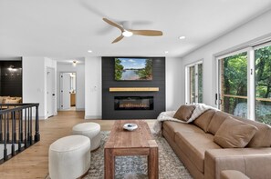 A modern living room with a fireplace, a ceiling fan, and a large window. - Sunset Cove — Lake Keowee Luxury Retreat with Private Dock + Hot Tub (Seneca)