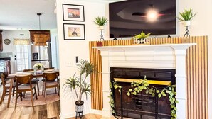 Ein Wohnzimmer mit Kamin, einem darüber montierten Fernseher und einem Essbereich mit Tisch und Stühlen.