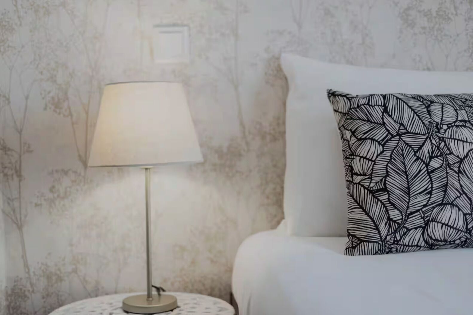 Un lit avec un oreiller blanc et un autre à motifs, une table de chevet avec une lampe, et un mur orné d’un motif discret représentant des feuilles.