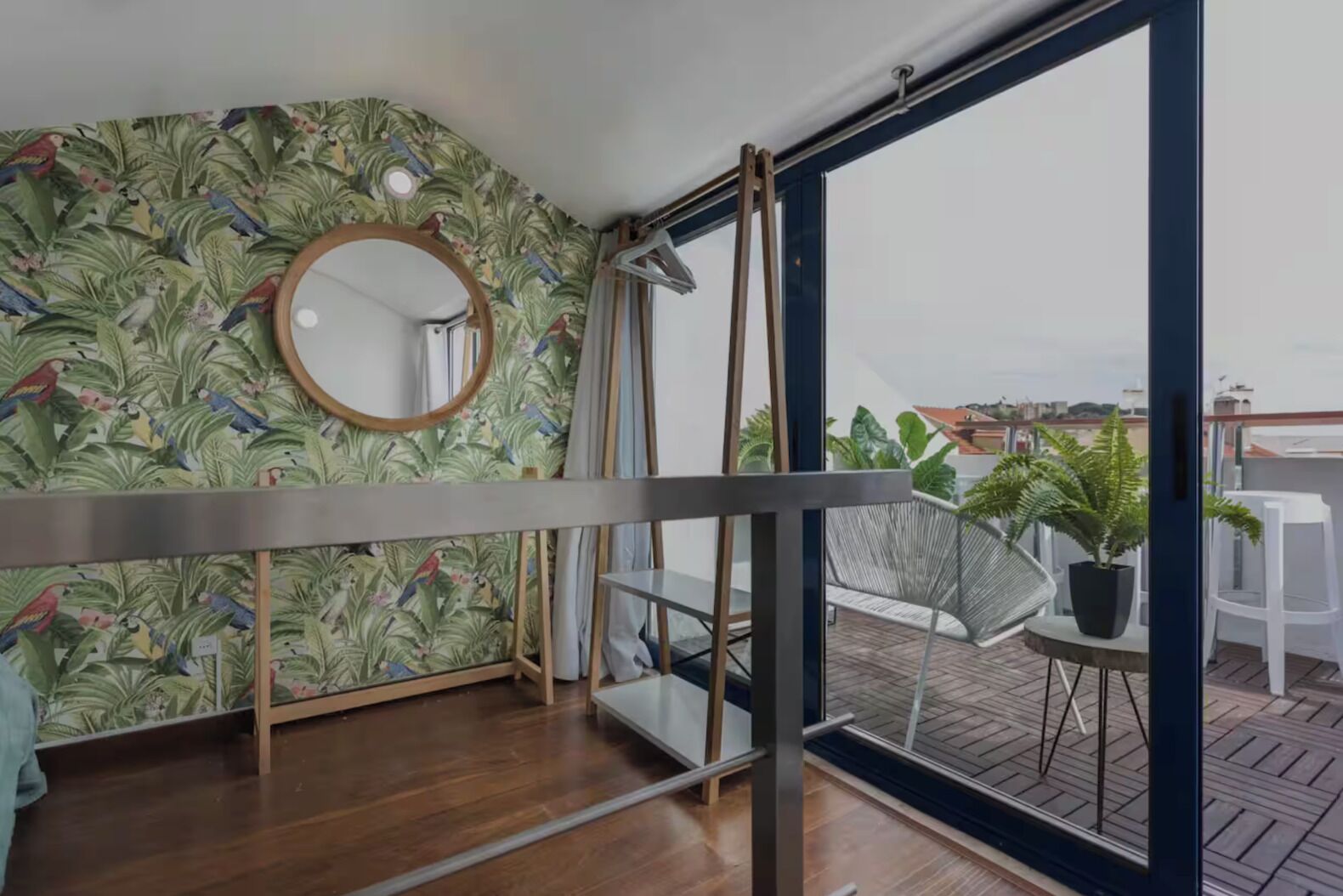 A balcony with a wooden floor, a metal railing, a round mirror, and a view of the city.