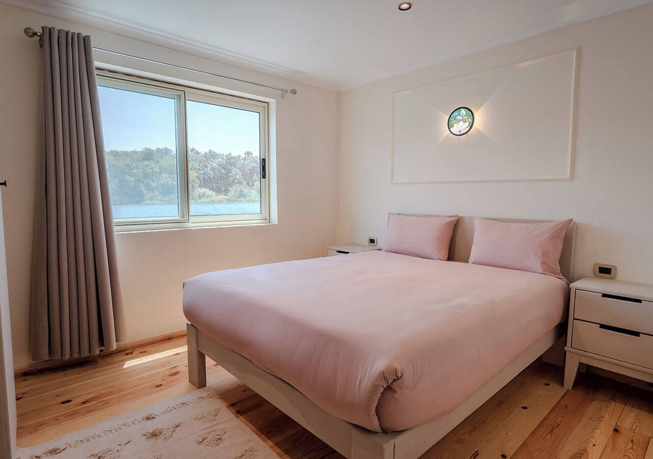 A bedroom with a bed, a nightstand, a window with curtains, and a wall-mounted light fixture.
