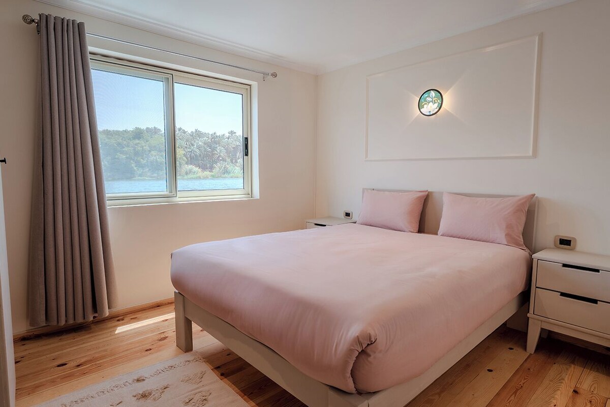 Une chambre à coucher comprenant un lit, une table de chevet, une fenêtre avec des rideaux et un luminaire fixé au mur.