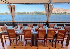 Un espace repas avec des tables et des chaises en bois, situé devant un paysage composé d’une rivière et de collines lointaines.