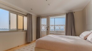 A bedroom with a large bed, wooden flooring, a view through a sliding door, and a window with curtains. - Prince Anoush Dahabiya 3 Nights (Luxor)