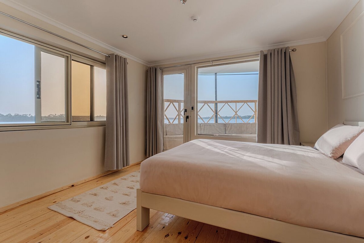 Une chambre avec un grand lit, un plancher en bois, une vue à travers une porte coulissante et une fenêtre avec des rideaux.