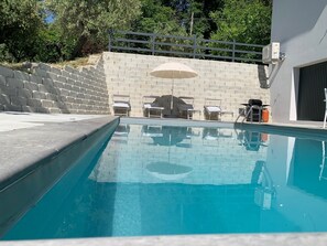 Ein Swimmingpool mit glasklarer, blauer Wasserfläche, umgeben von einer niedrigen Steinmauer, ausgestattet mit Liegestühlen und einem Sonnenschirm.