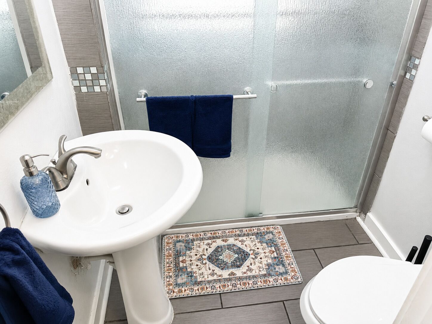 Un baño con lavamanos blanco, cabina de ducha de vidrio, toalla azul y alfombra estampada.