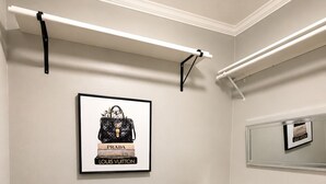 Et værelse med et sort skrivebord, en grå stol, et indrammet billede af designertasker og et spejl på væggen.