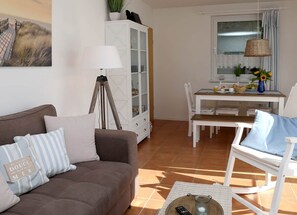 A living room with a brown sofa, a white cabinet, a dining table with chairs, and a window with curtains.