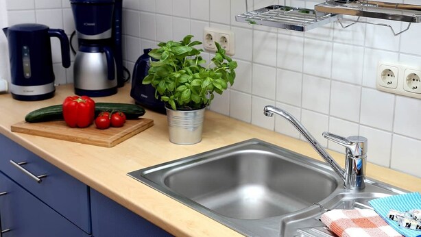 Una cocina con un lavamanos de acero inoxidable, una cafetera, una planta de albahaca en maceta y una tabla de cortar con verduras.