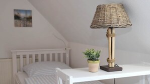 Un dormitorio con una cama blanca, una mesita de noche con una lámpara y una planta en maceta.