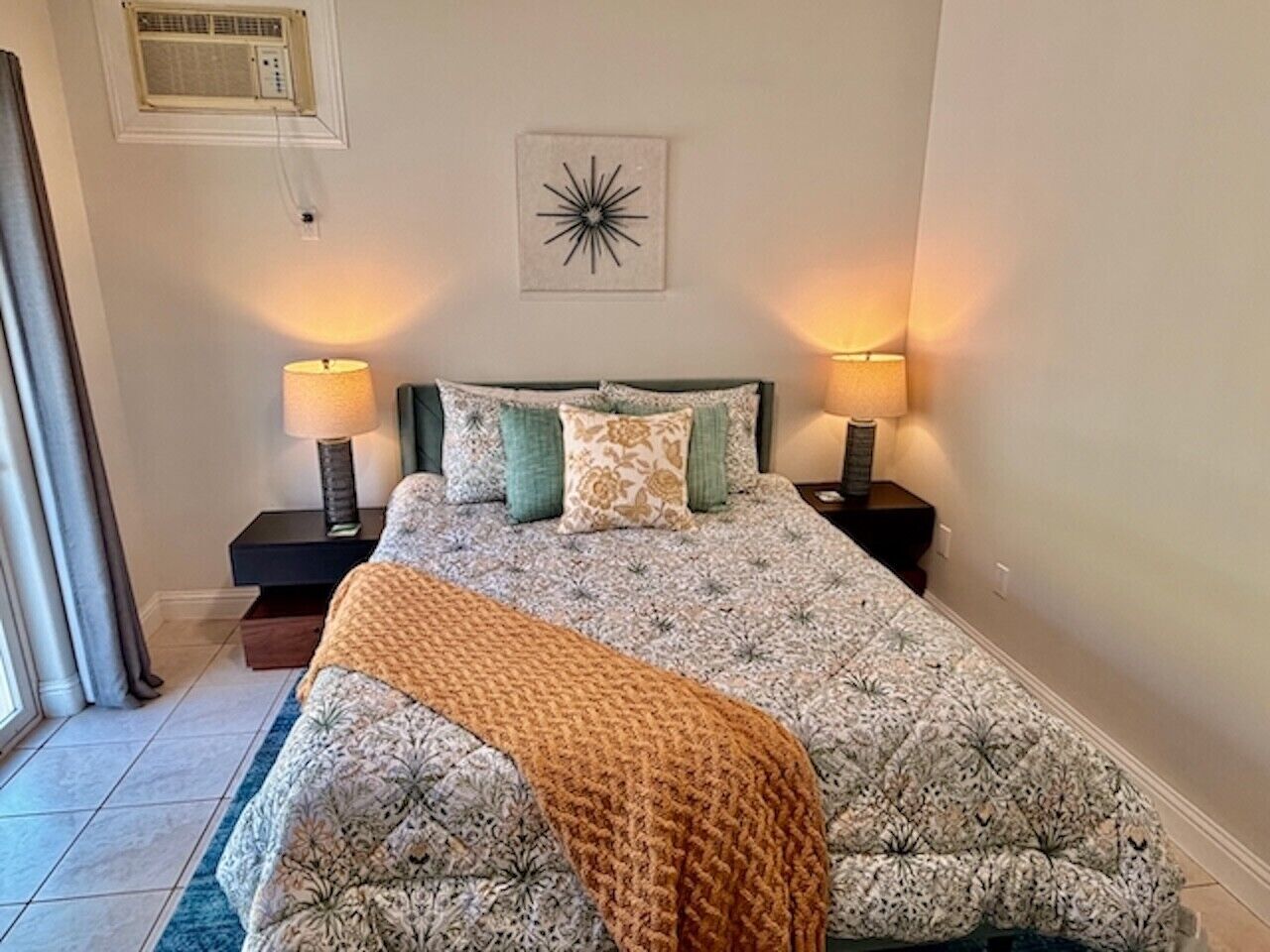 Quarto com cama, duas mesas de cabeceira, uma luminária sobre cada mesa, um detalhe decorativo na parede e uma janela com cortinas.