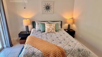 Quarto com cama, duas mesas de cabeceira, uma luminária sobre cada mesa, um detalhe decorativo na parede e uma janela com cortinas.
