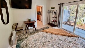 Quarto com cama, escrivaninha, cadeira, televisão, ventoinha no teto e vista para o exterior através de uma porta de correr.