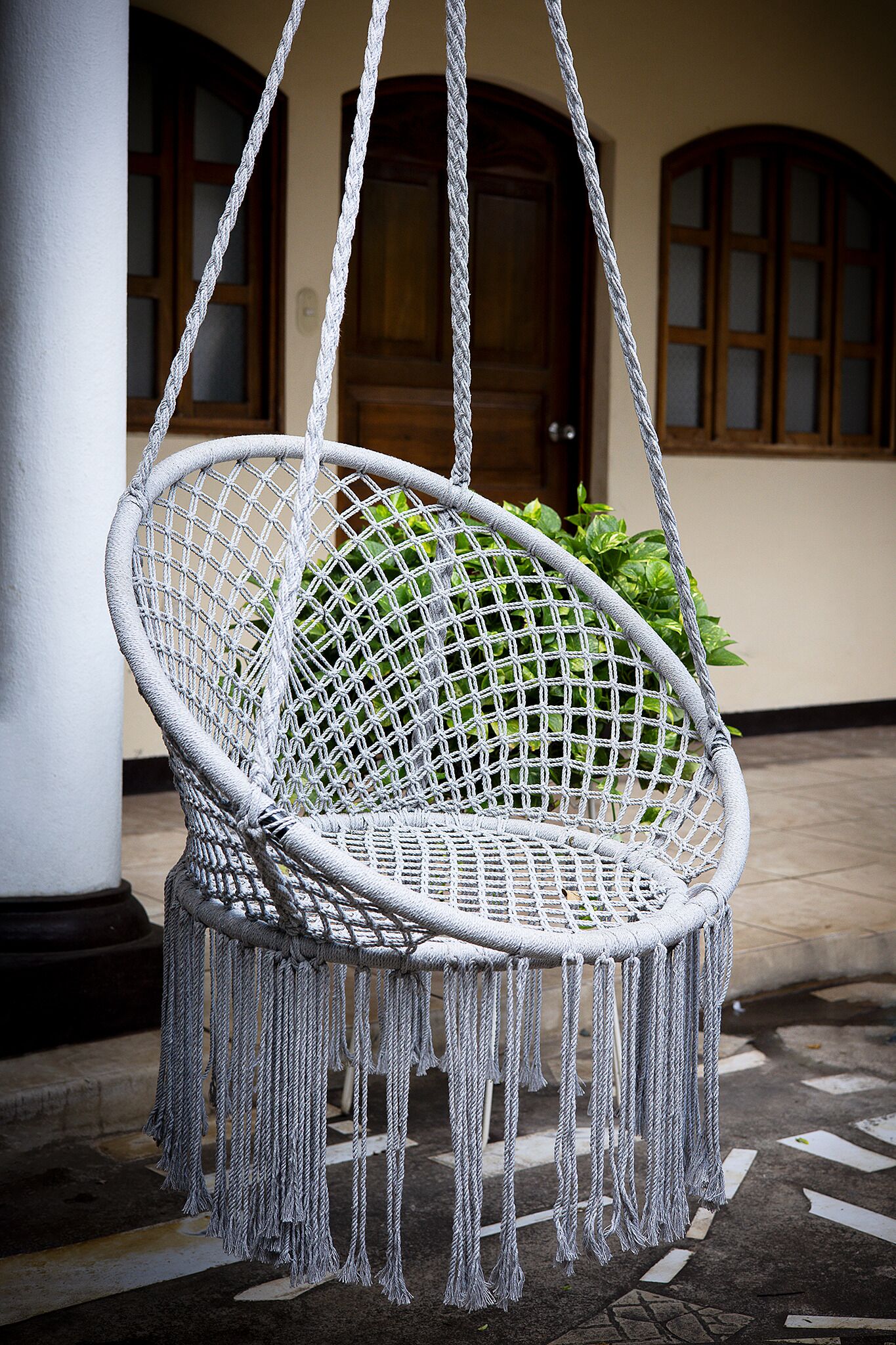 Un fauteuil suspendu en macramé blanc, orné de franges, maintenu par des cordes, devant un bâtiment doté de portes et de fenêtres en bois.