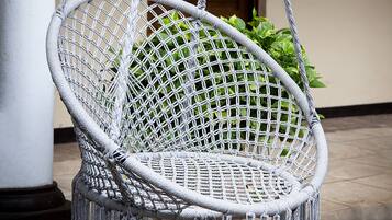 Un fauteuil suspendu en macramé blanc, orné de franges, maintenu par des cordes, devant un bâtiment doté de portes et de fenêtres en bois.