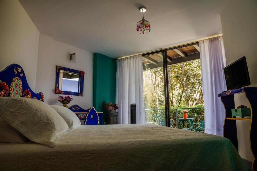 Une chambre à coucher avec un lit, un miroir, une télévision et un balcon donnant sur la forêt.