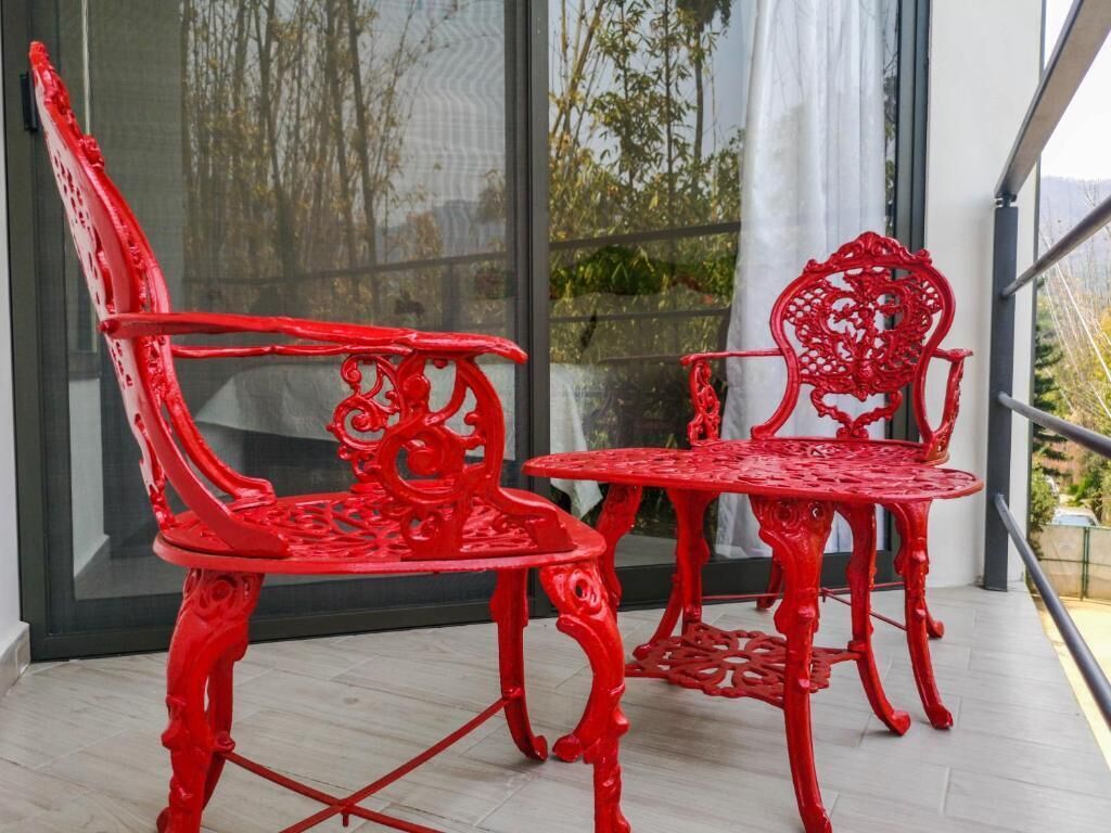 Two red chairs and a small table on a balcony with a view of trees and a building.